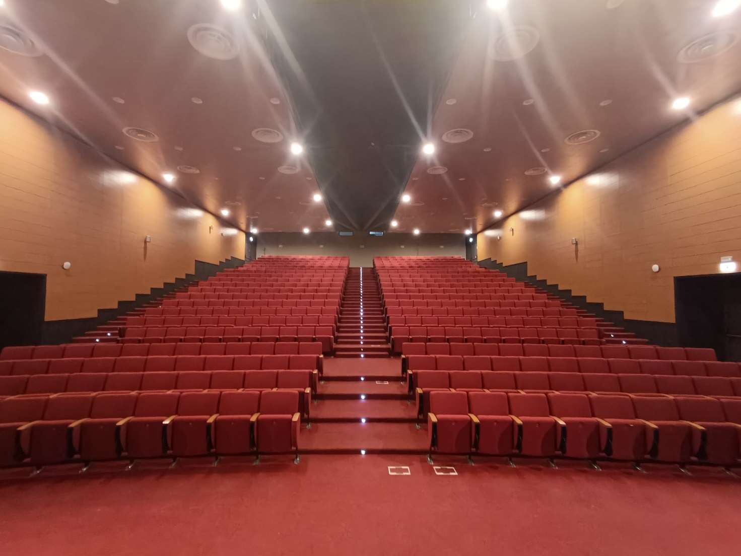 View down the centre aisle of the Aula Magna CTO auditorium showing tiered rows of red seats and timber-panelled walls